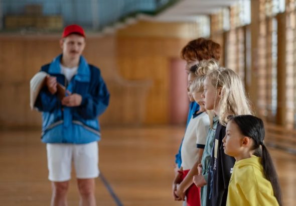 Diverse group of children standing indoors in a gym with a coach in focus.